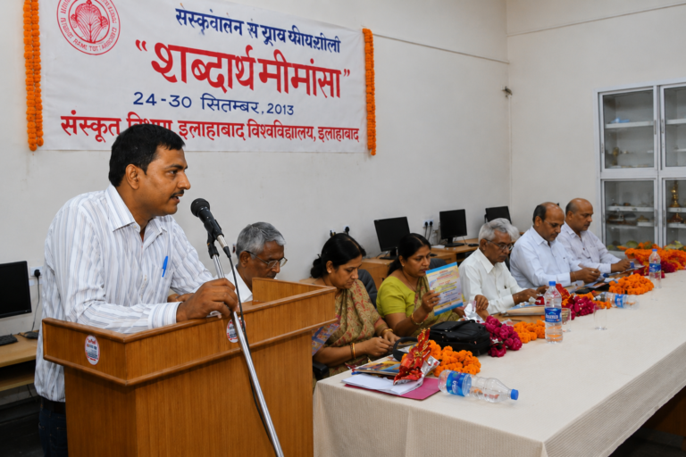 A speaker standing at a wooden podium and addressing a panel during a Sanskrit academic seminar in a conference hall.
