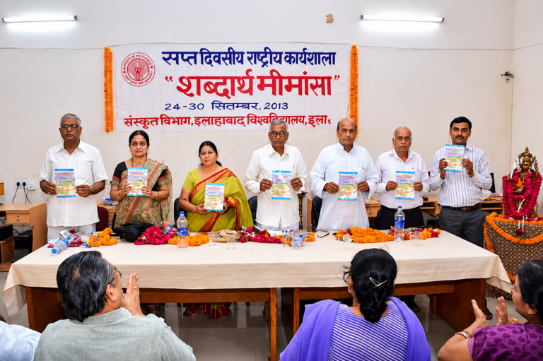Wide indoor conference scene with scholars holding seminar booklets while attendees applaud during an academic Sanskrit event.